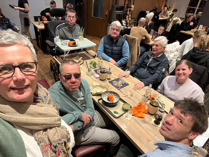 sechs Barrierecheckende sitzen gemeinsam an einem Tisch, Frau Wollenweber macht ein Selfie der Gruppe Das Bild zeigt die Gruppe der Barrierecheckende die um einen Tisch herum sitzen