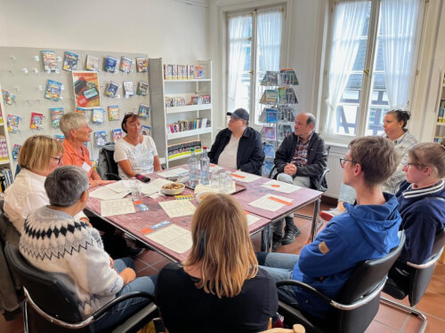Neun Personen sitzen um einen mit Papieren und Wasserflaschen bedeckten Tisch herum und diskutieren in einem hell erleuchteten Raum mit Regalen voller Zeitschriften und B&uuml;cher im Hintergrund.