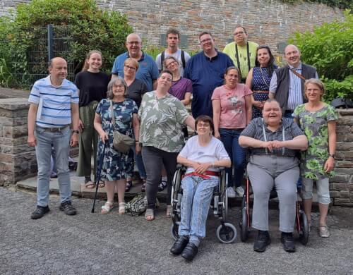 20250617_194131 Eine Gruppe von fünfzehn Erwachsenen, darunter zwei Rollstuhlfahrer, posieren gemeinsam vor einer Steinmauer und einer Grünanlage.