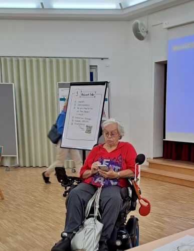 Eine &auml;ltere Frau in einem motorisierten Rollstuhl benutzt ihr Smartphone in einem Besprechungsraum mit einem Pr&auml;sentationsbildschirm und einem Flipchart im Hintergrund.