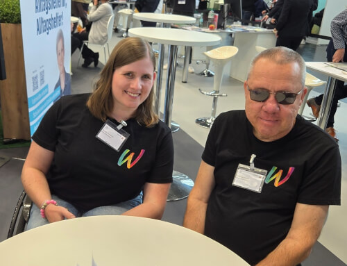 Bodo und Pauline Zwei Personen, die passende schwarze T-Shirts mit einem bunten W tragen, sitzen an einem runden Tisch in einem Veranstaltungsraum mit anderen Teilnehmern im Hintergrund.