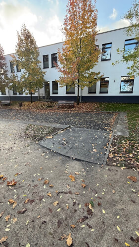 Eine Rollstuhlrampe f&uuml;hrt zu einem erh&ouml;hten Weg vor einem wei&szlig;en Geb&auml;ude mit gro&szlig;en Fenstern und Herbstb&auml;umen. Gefallene Bl&auml;tter bedecken den Boden.