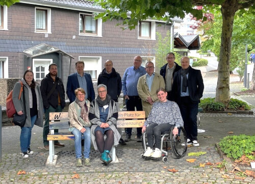 Gruppenfoto Schulterblick Eine Gruppe von elf Erwachsenen, darunter eine Person im Rollstuhl, posiert im Freien neben einer Bank und einem Schild auf einem Kopfsteinpflaster vor Wohnhäusern.