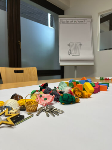 Auf einem Tisch in einem Besprechungsraum sind verschiedene kleine Gegenst&auml;nde und bunte Bl&ouml;cke angeordnet, w&auml;hrend im Hintergrund ein Flipchart mit der Zeichnung eines M&uuml;lleimers zu sehen ist.