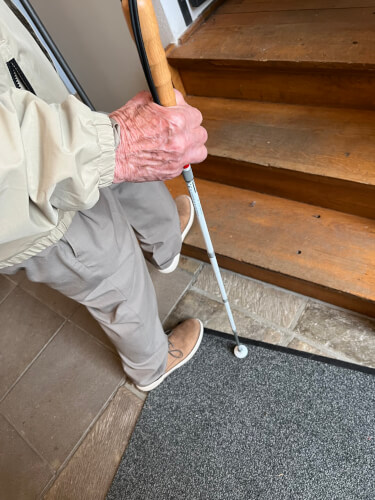 image00012 Eine ältere Person mit einem weißen Stock steht am Fuß einer Holztreppe und trägt eine beigefarbene Hose, eine helle Jacke und hellbraune Schuhe.