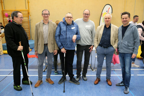 Sechs M&auml;nner stehen in einer Turnhalle zusammen, einige mit St&ouml;cken, und posieren f&uuml;r ein Gruppenfoto. Im Hintergrund sind St&uuml;hle, Turnger&auml;te und andere Personen zu sehen.