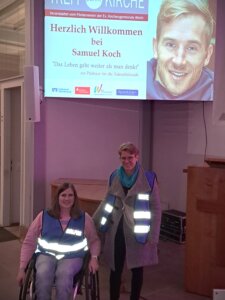 Zwei Frauen mit Warnwesten, eine davon im Rollstuhl, stehen in einem Innenraum. Hinter ihnen ist ein großes projiziertes Willkommensschild für eine Samuel-Koch-Veranstaltung zu sehen.