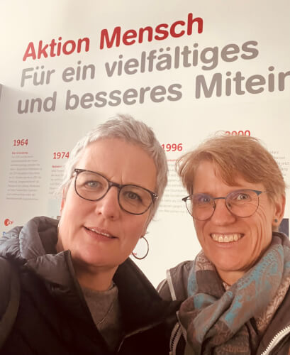 image00001 Zwei Frauen mit kurzen Haaren und Brillen posieren vor einer Wand mit deutschem Text über die Aktion Mensch und die Förderung von Vielfalt und Zusammengehörigkeit.
