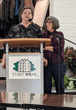 Bild1 Zwei Frauen stehen an einem Podium mit einem Schild der Stadt Wiehl; eine spricht in ein Mikrofon, während die andere neben ihr steht und eine Hand auf ihren Arm legt. Im Hintergrund sind Mäntel und Blumen zu sehen.