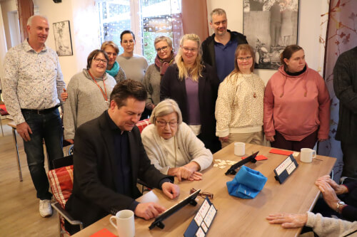 Eine Gruppe von Menschen, darunter Senioren und Erwachsene, versammelt sich in einem Innenraum um einen Tisch und beobachtet einen Mann, der ein Tablet benutzt, w&auml;hrend andere ihn beobachten und l&auml;cheln.