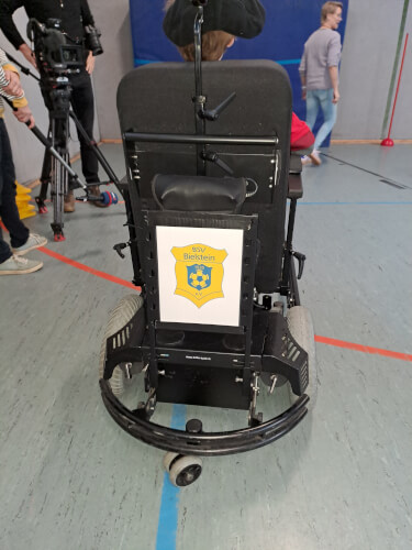 20251213_140344 Ein Elektrorollstuhl mit einem BSV Bielstein-Schriftzug auf der Rückseite ist in einer Turnhalle geparkt, im Hintergrund sind Menschen und eine Kamera zu sehen.