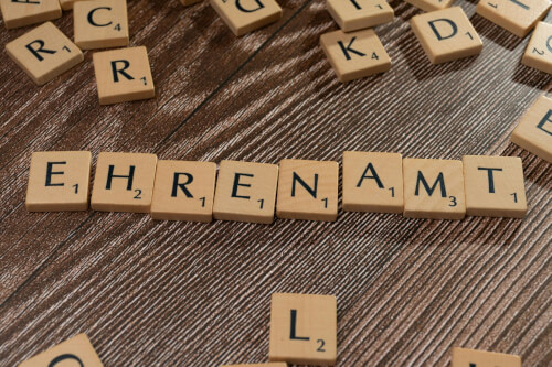 Auf einem Tisch liegen h&ouml;lzerne Buchstabenpl&auml;ttchen, die das deutsche Wort EHRENAMT buchstabieren, umgeben von anderen verstreuten Buchstabenpl&auml;ttchen.