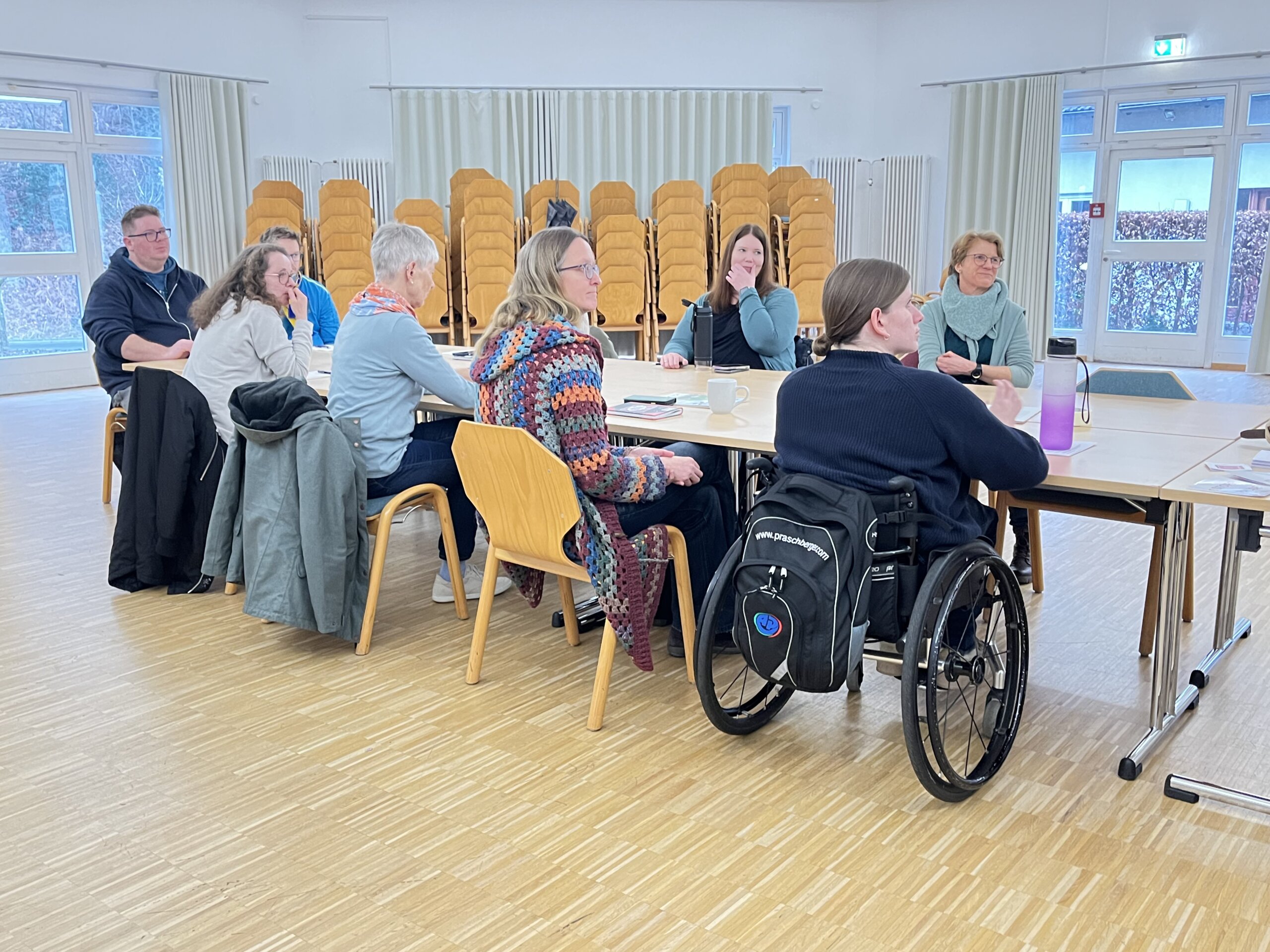 9 personen sitzen um einen tisch und h&ouml;ren einer sprecherin zu