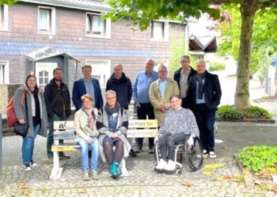 Eine Gruppe von elf Erwachsenen, darunter eine Person im Rollstuhl, posiert gemeinsam im Freien in der Nähe einer Bank und eines Baumes vor einem Wohngebäude.