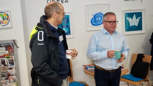 Zwei Männer stehen und unterhalten sich, während sie Tassen in einem Innenraum halten. Im Hintergrund sind gerahmte Kunstwerke und Stühle zu sehen.