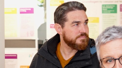 Ein Mann mit kurzen braunen Haaren und einem Bart, der ein braunes Hemd und eine dunkle Jacke trägt, steht in einem Innenraum vor einer Wand mit bunten Postern.