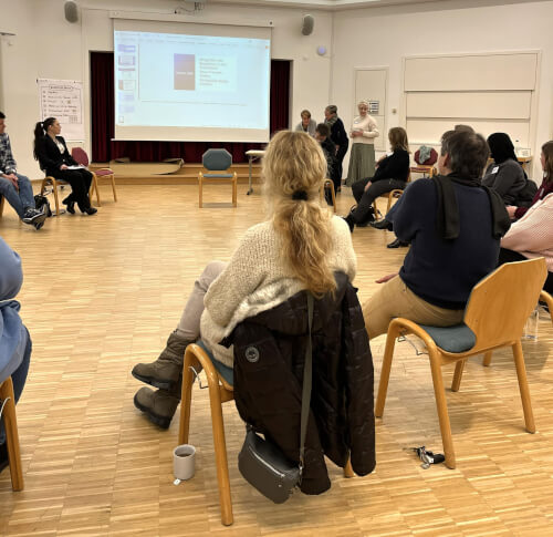 Netzwerk Gruppe 2 Eine Gruppe von Personen sitzt in einem Halbkreis in einem Besprechungsraum und blickt auf eine projizierte Präsentationsleinwand und die vorne stehenden Redner.