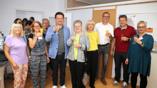 Eine Gruppe von Menschen steht in einem Haus, lächelt in die Kamera und hält Getränke in der Hand, als ob sie feiern oder an einem gesellschaftlichen Ereignis teilnehmen würden.