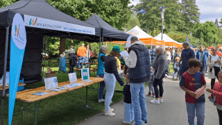 An einem sonnigen Tag versammeln sich Menschen an Informationsständen mit Bannern und Tischen mit Broschüren in einem Park.