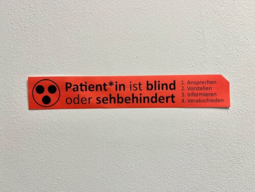 Bild Aufkleber 1 Rotes Schild an einer Wand mit deutschem Text, der angibt, dass der Patient blind oder sehbehindert ist, und einer Liste mit vier Schritten, die das Personal befolgen soll.