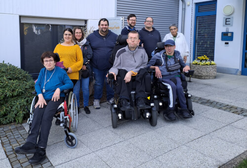 Eine Gruppe von neun Erwachsenen, darunter drei Rollstuhlfahrer, posiert gemeinsam vor einem Geb&auml;ude mit einer blauen T&uuml;r und Topfblumen.