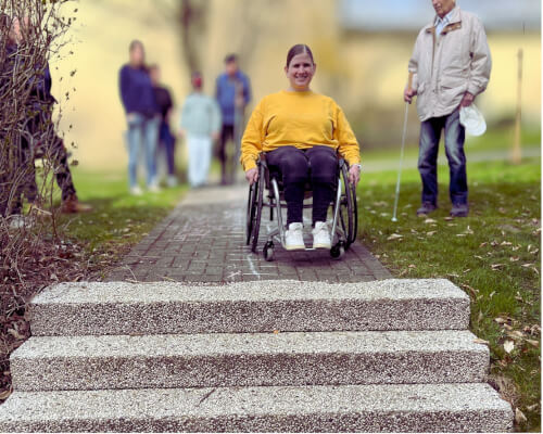 Eine Frau im Rollstuhl steht vor einer Treppe auf einem Gehweg, während im Hintergrund mehrere Personen stehen.