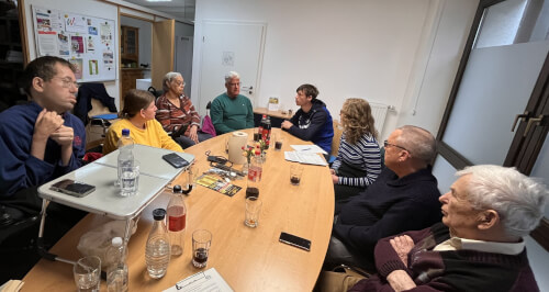 Acht Personen sitzen in einem hell erleuchteten Besprechungsraum an einem Konferenztisch und diskutieren, während Getränke, Unterlagen und Snacks auf dem Tisch stehen.