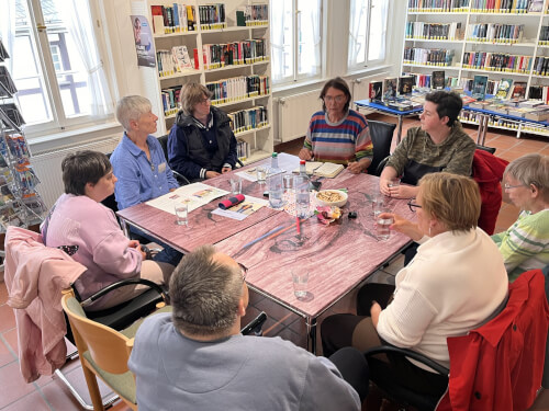 Eine Gruppe von acht Erwachsenen sitzt um einen Tisch mit Büchern und Getränken und diskutiert in einer Bibliothek.