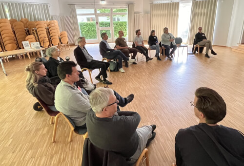 Eine Gruppe von Menschen sitzt im Kreis auf Stühlen in einem hellen Raum mit gestapelten Stühlen und großen Fenstern im Hintergrund.
